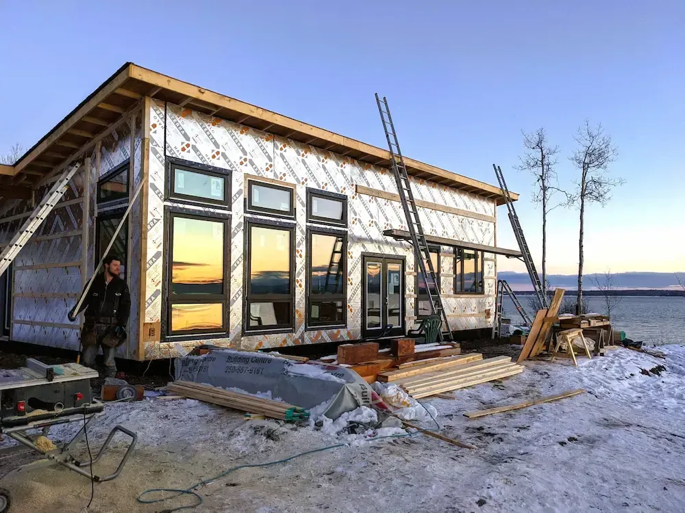 House under construction with large windows on a snowy shoreline at sunset