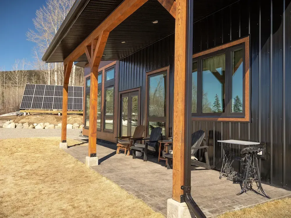 Modern house porch with wooden posts, large windows, black siding, and patio chairs on a concrete patio.