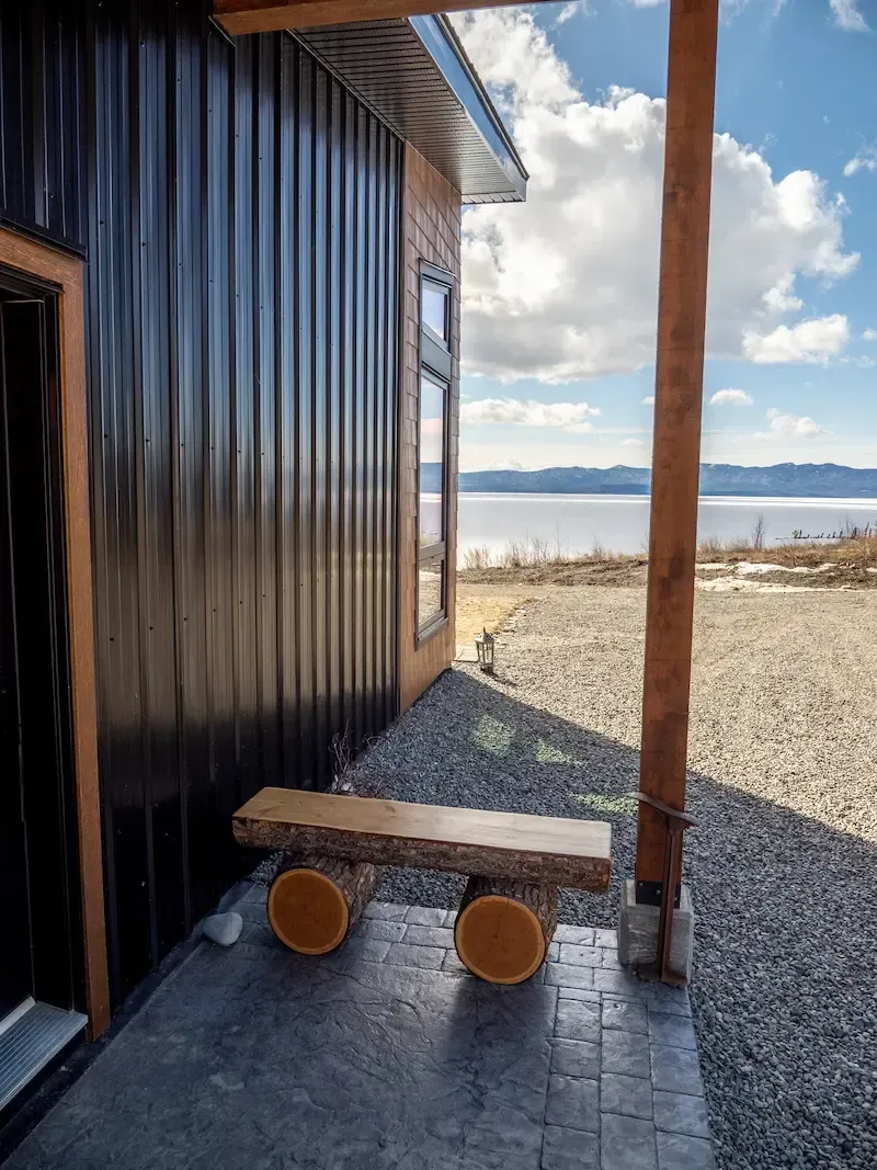 Black cabin exterior with a small wooden cart on a gravel path beside a snowy waterfront