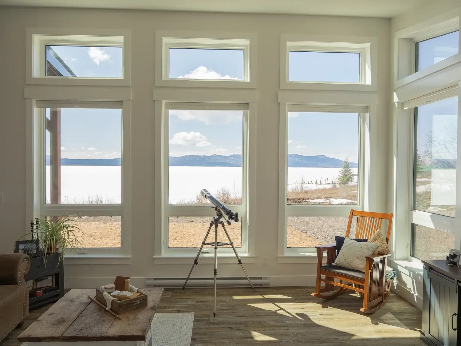 Bright living room with large windows, telescope, chair, and a view of water and mountains.