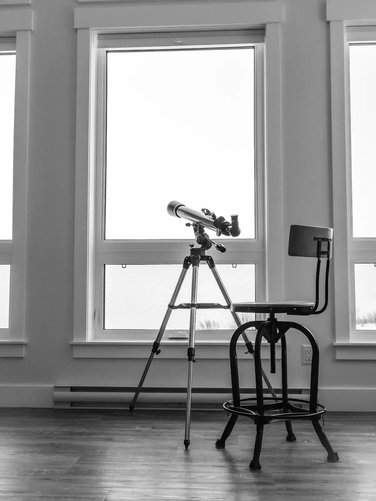 Telescope on a tripod by a window, with a stool and wooden floor in a bright room.