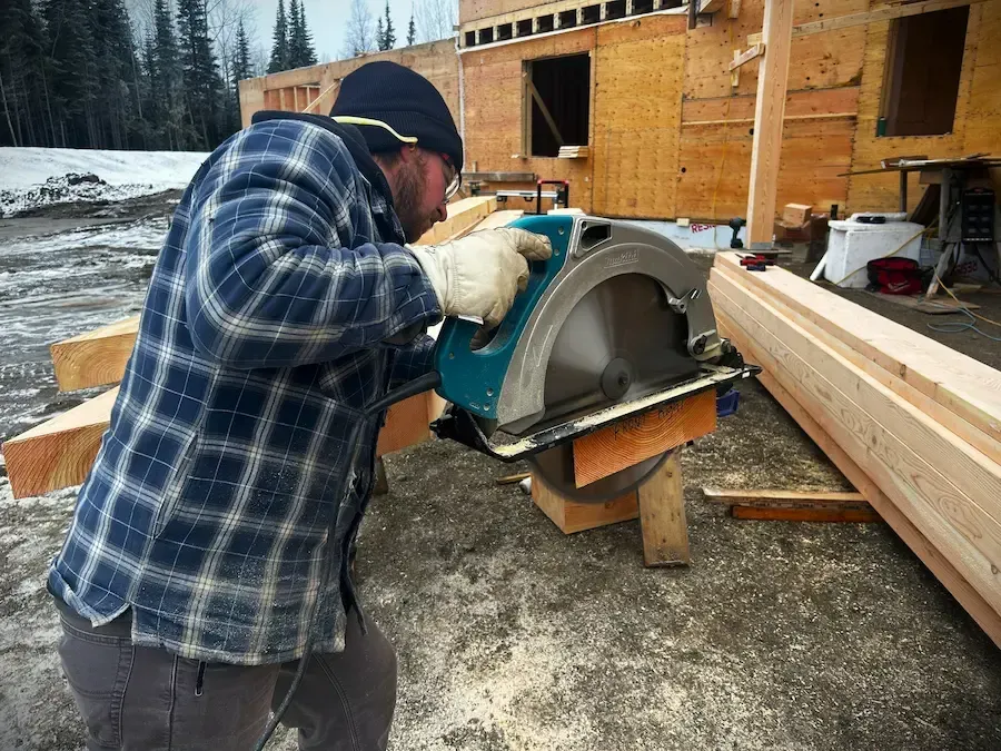 Worker cutting a metal piece outdoors beside lumber and a wooden building site