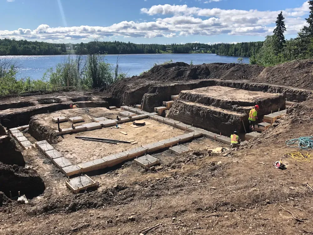 Archaeological excavation beside a lake, with stone foundations and two workers in high-visibility vests.