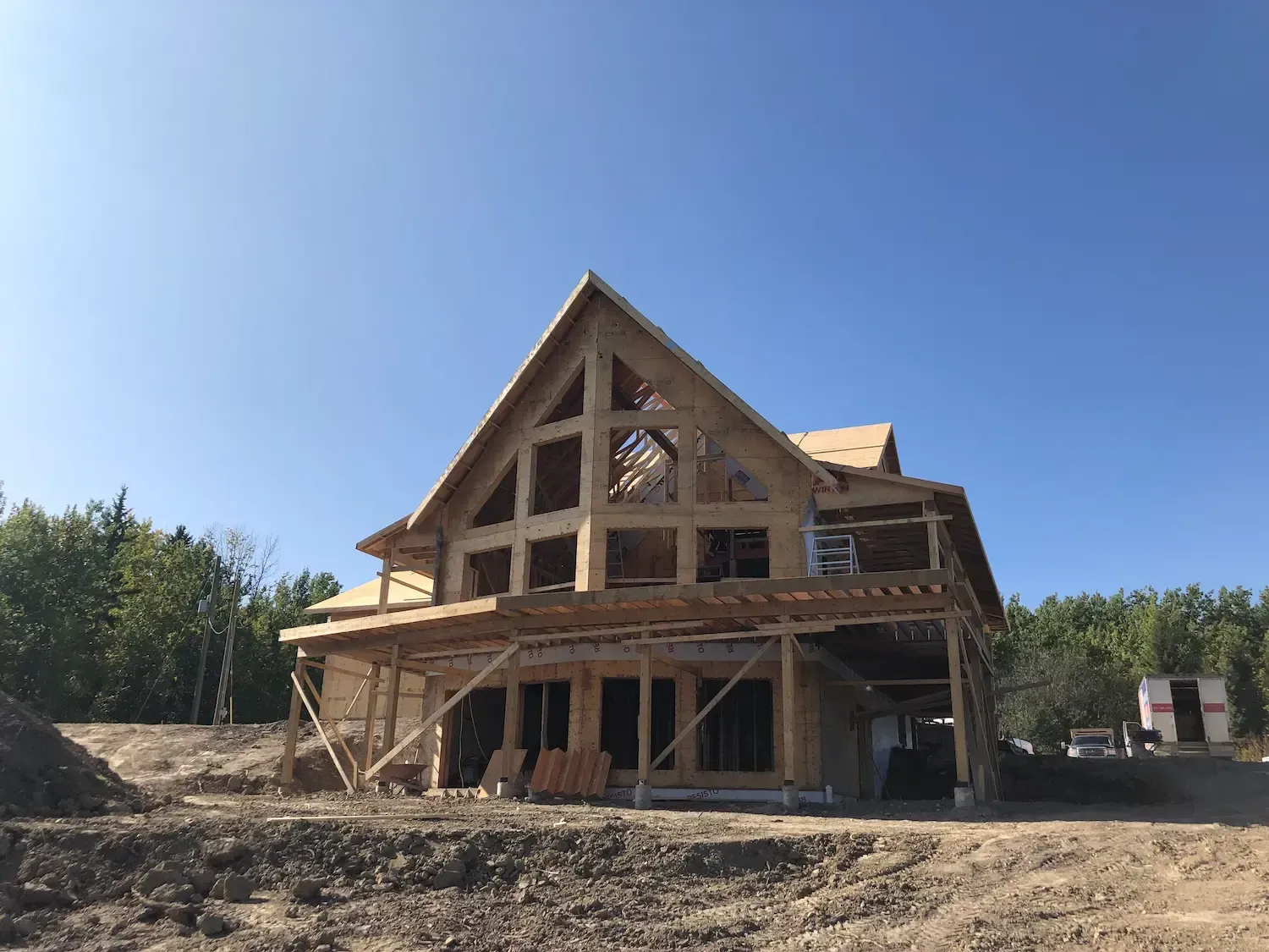 Partially framed wooden house under construction on a dirt lot under a clear blue sky
