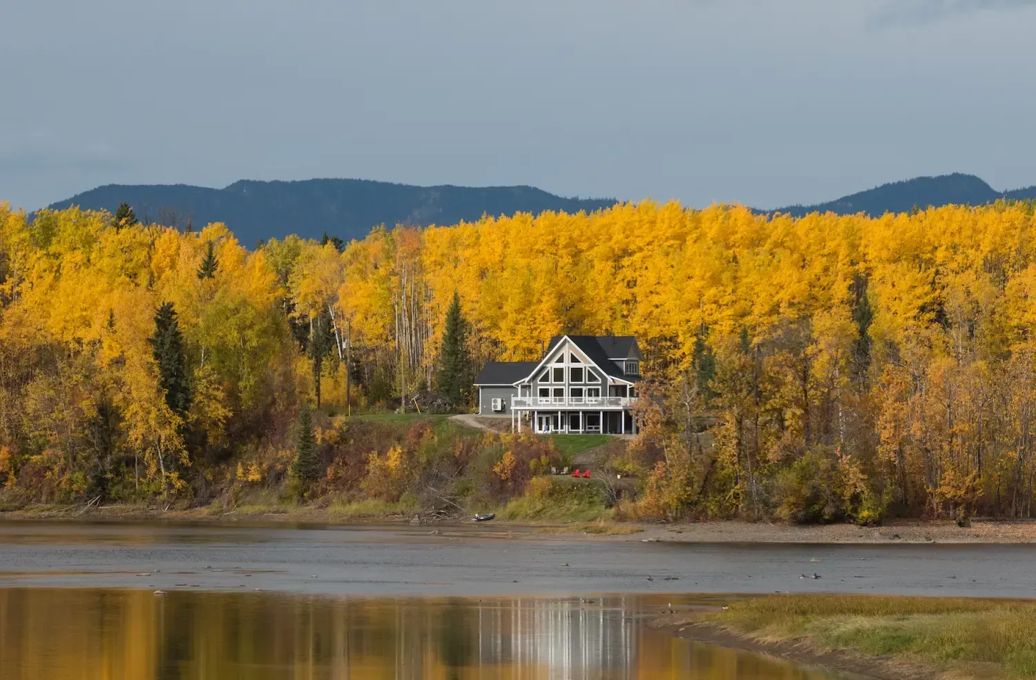 Lakeside house backed by golden autumn trees and distant blue hills, reflected in calm water