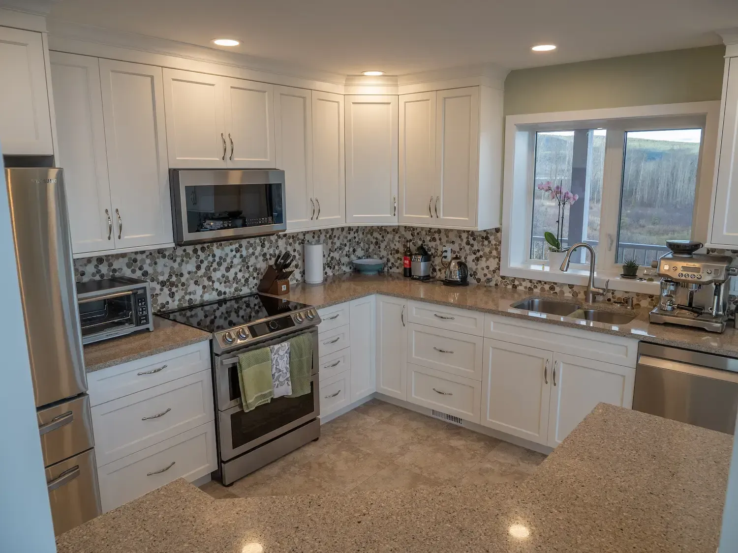 Bright white kitchen with stainless appliances, speckled granite counters, and a large window overlooking a patio.