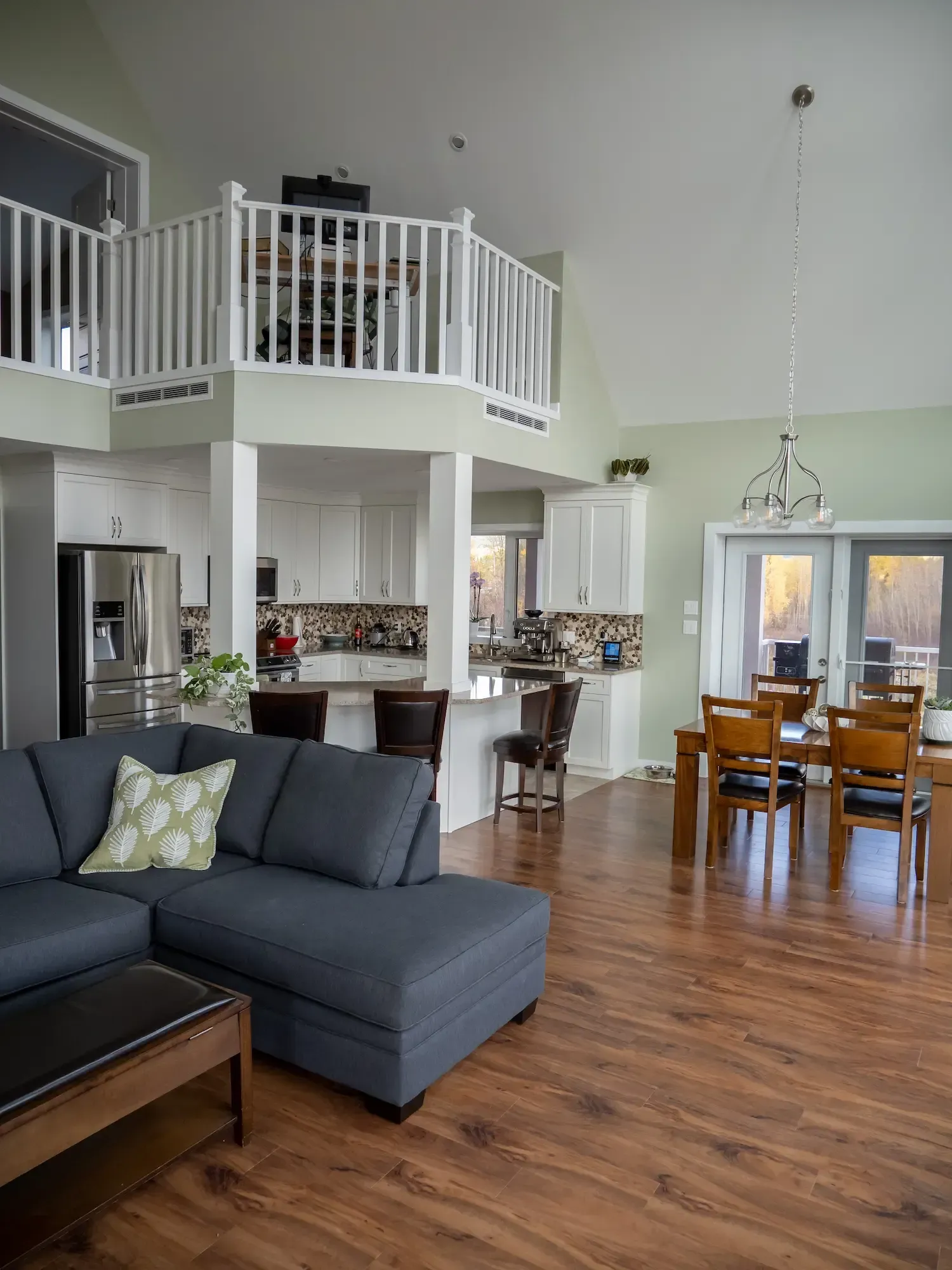 Open-plan living room with gray sectional, wood floors, loft railing, and dining area near French doors