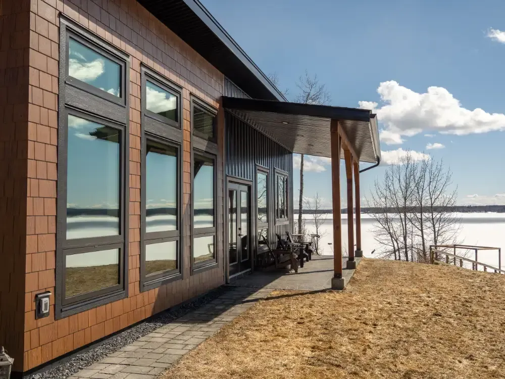 Modern house exterior with wood siding, large windows, and a covered patio overlooking a snowy landscape