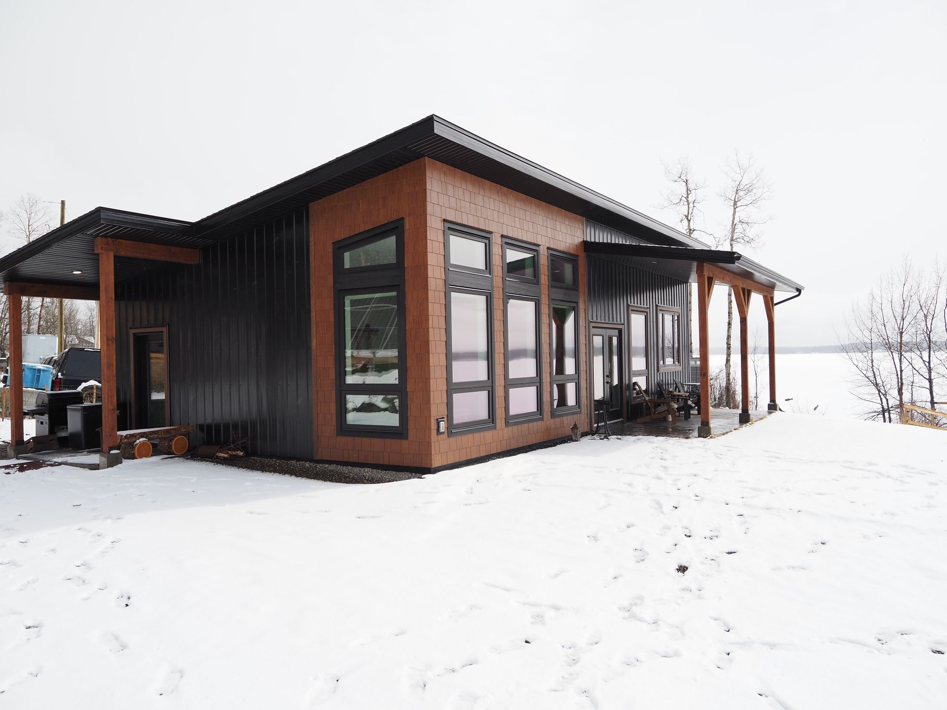 Modern cabin with dark wood siding and large windows, sitting in a snowy landscape.