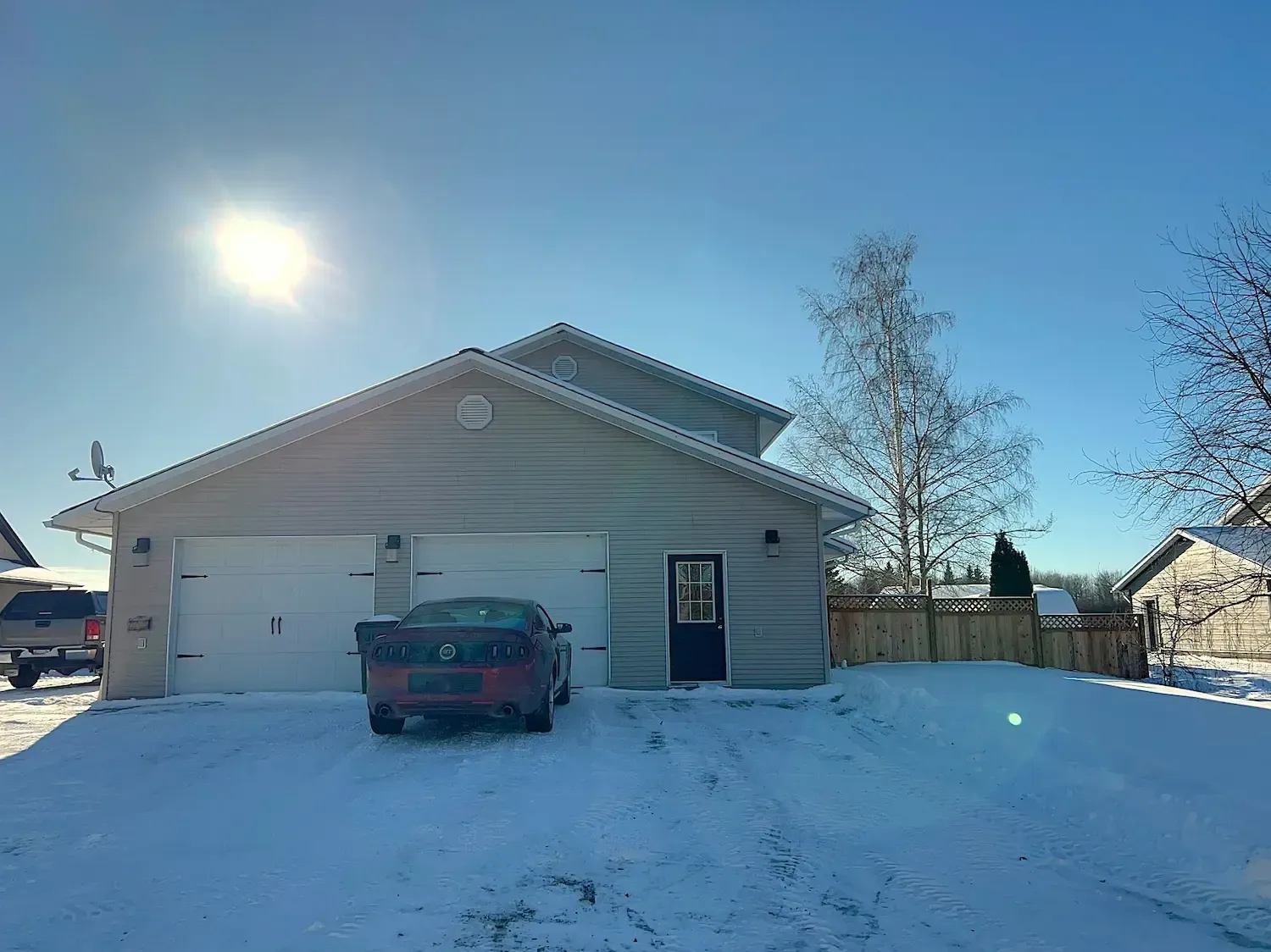 Snowy suburban house with a red car in the driveway and bright sun overhead