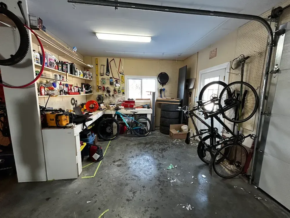 Cluttered garage with tools, shelves, bicycles, and a workbench under a ceiling light