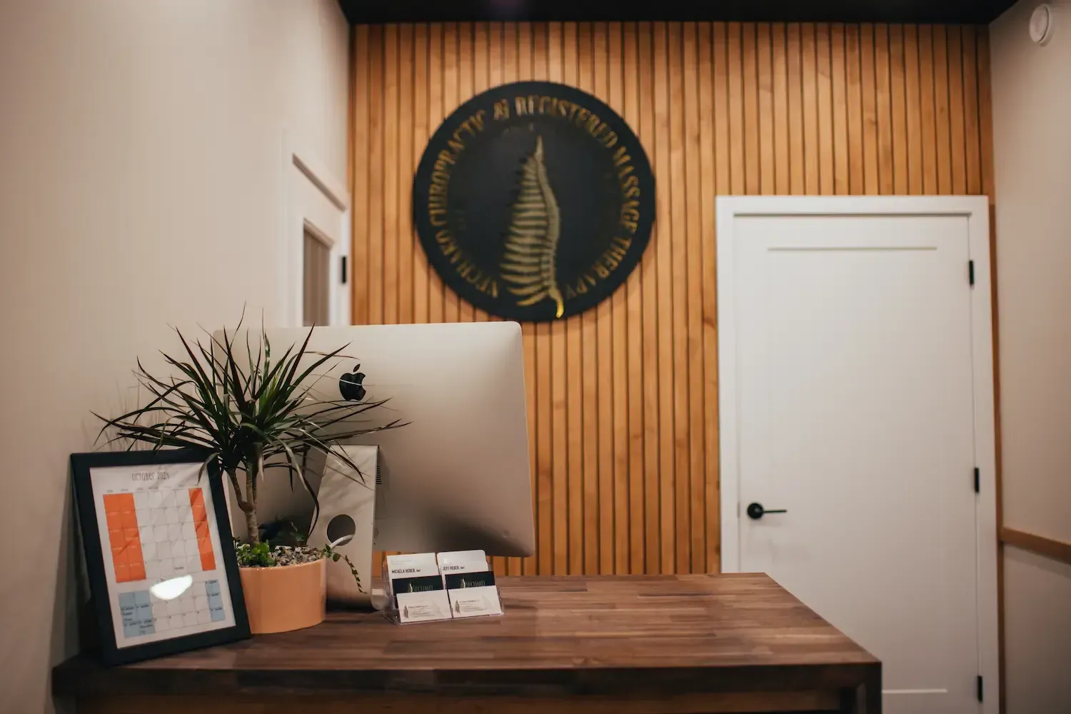 Wood-paneled reception area with a desk, potted plant, wall emblem, and white door.
