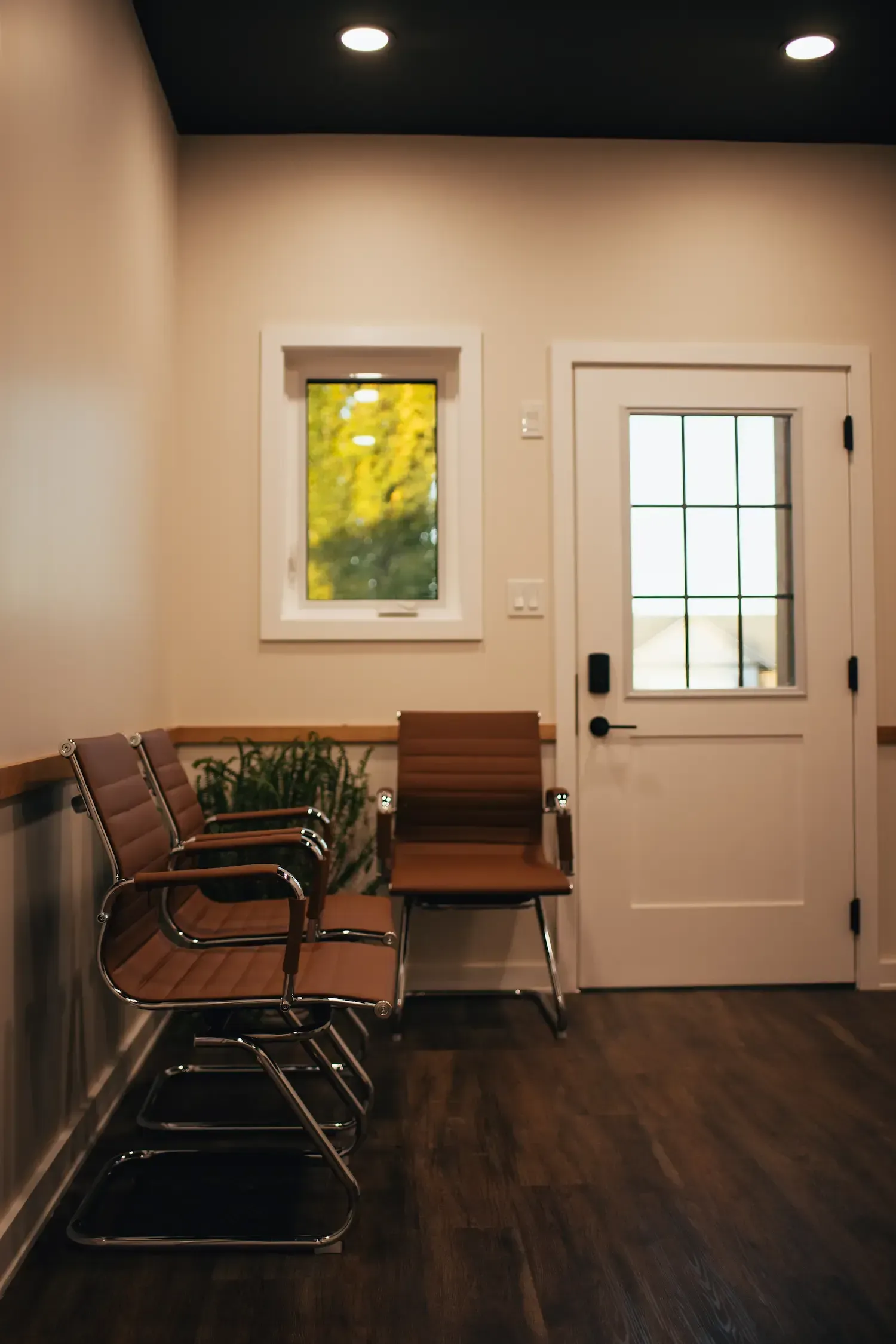 Quiet waiting room with stacked chairs, a small table, window, and white door on dark wood floor.