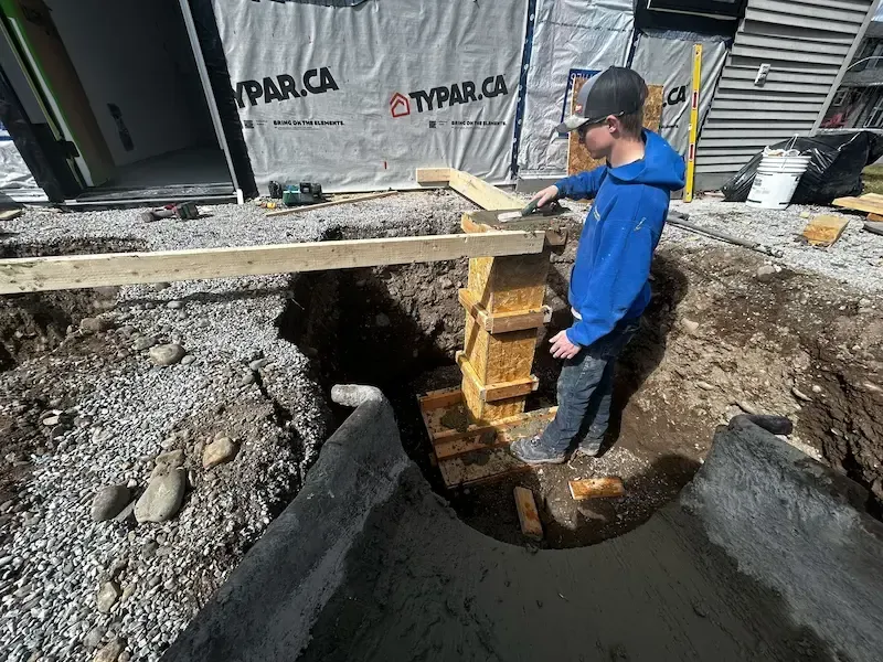 Worker in blue shirt stands beside a deep excavation, supporting a wooden beam at a construction site.