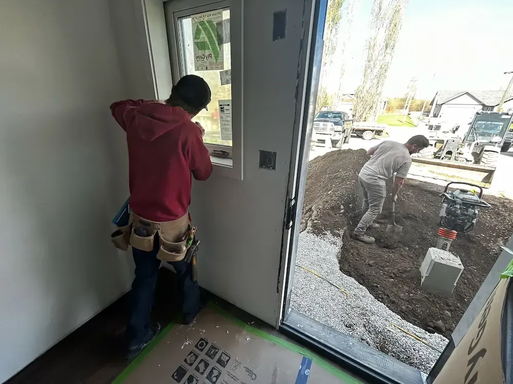 Worker inside house installing a door while another worker levels dirt outside