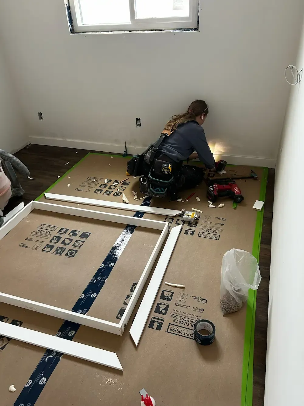 Person assembling a white frame on a floor-covered room with tools and supplies spread out nearby.