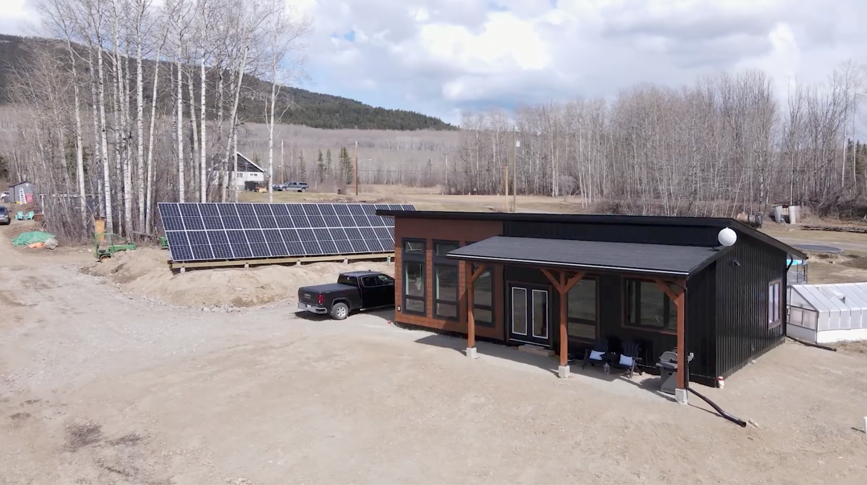 Small wooden cabin with solar panels in a rural clearing, surrounded by leafless trees and mountains.