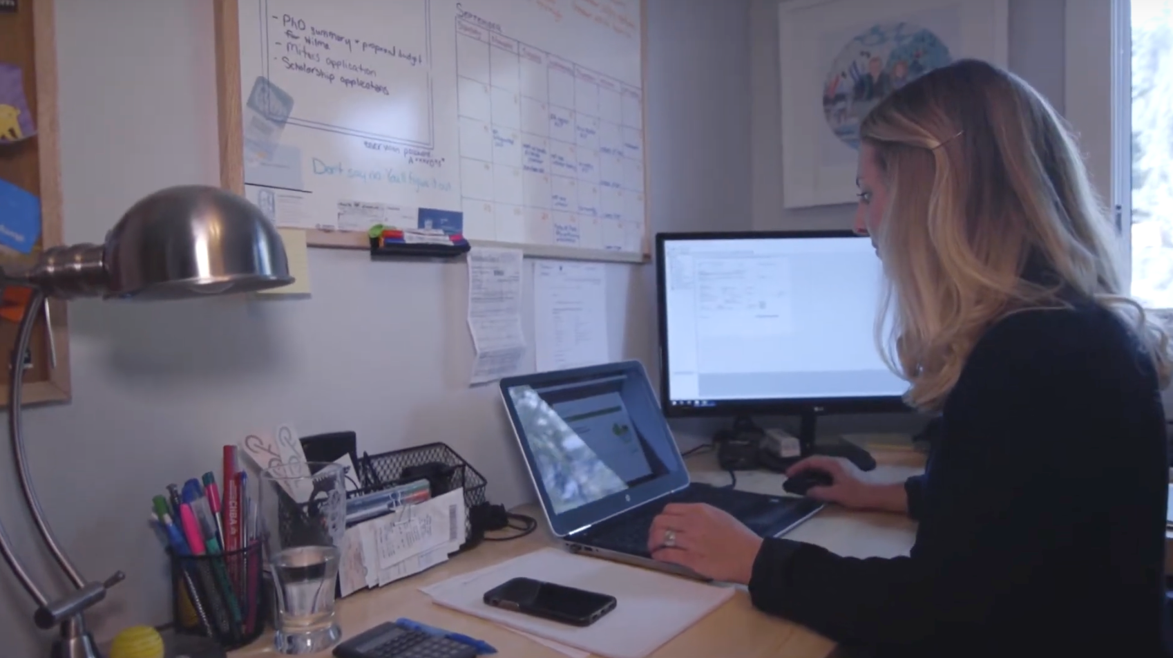 Person working at a desk with a laptop and monitor in a home office, papers and pens nearby.