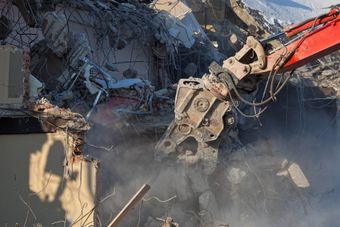 Un escavatore sta demolendo una struttura in cemento, sollevando polvere e detriti nell'aria.