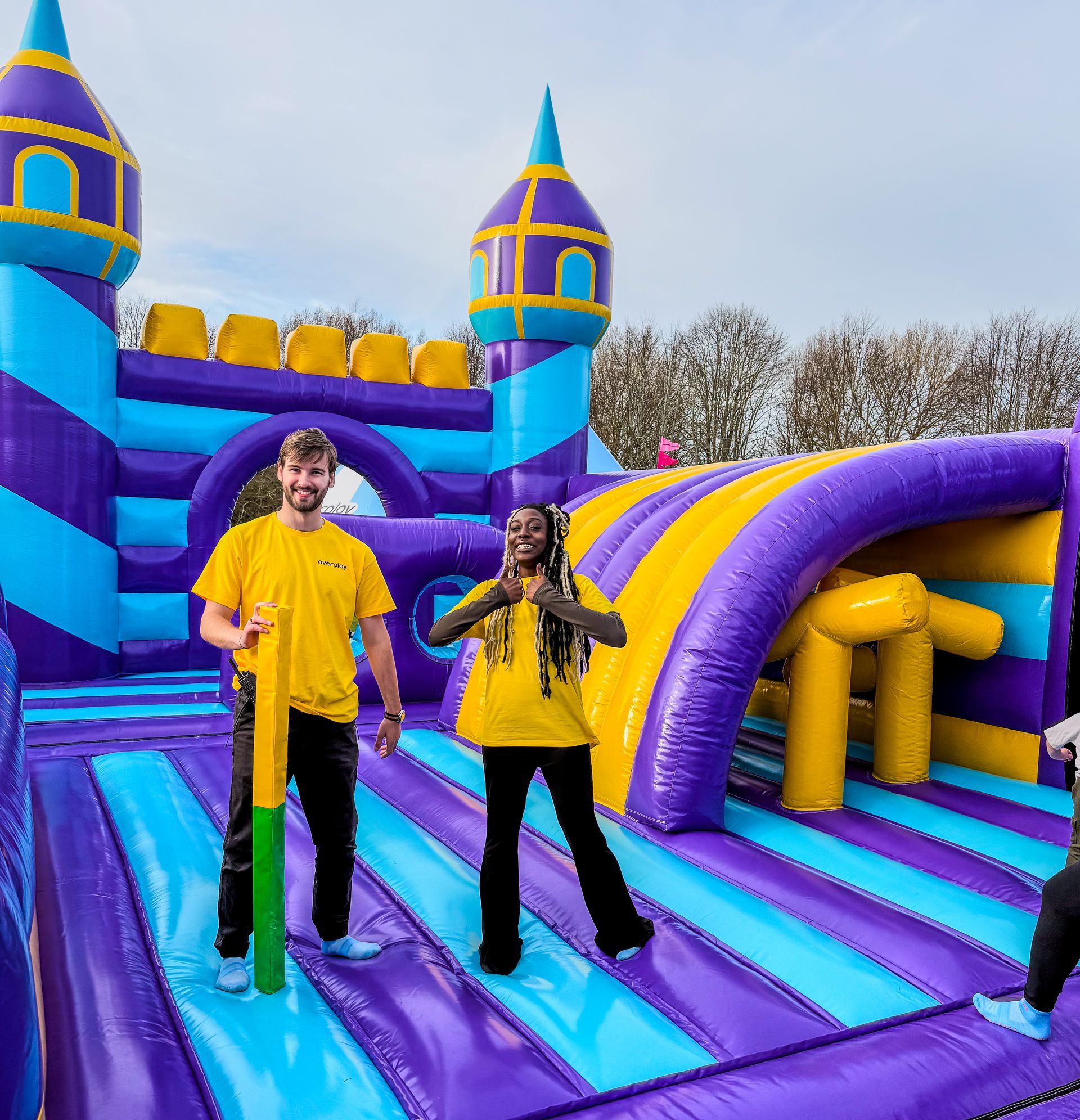 Two people in yellow shirts near a purple and blue inflatable castle. One holds a pole, the other gives a thumbs-up.