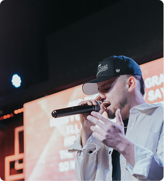 Singer in a black cap performing into a microphone on stage with warm orange lights
