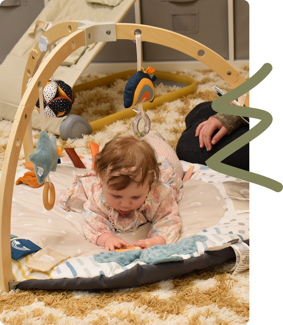 Baby crawling on a play mat under a wooden arch with hanging toys, next to an adult's hand.