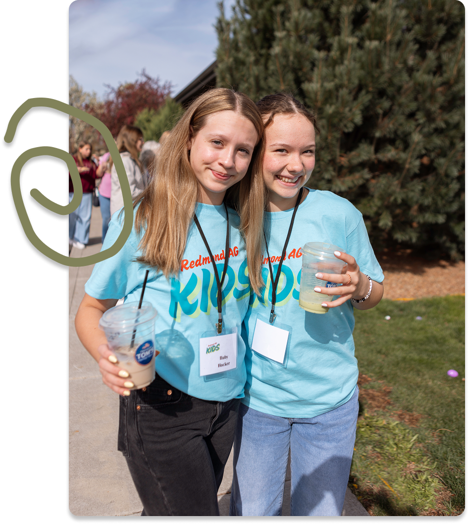 Two smiling girls in blue T-shirts holding drinks outdoors on a sunny day