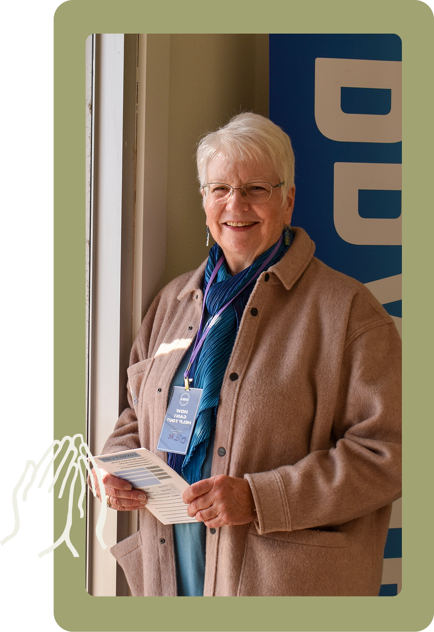 Smiling person in tan coat holding papers beside a doorway with a blue sign