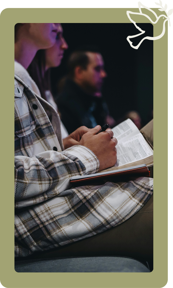 Person in a plaid shirt writes in a notebook while seated in a dim lecture or meeting room.