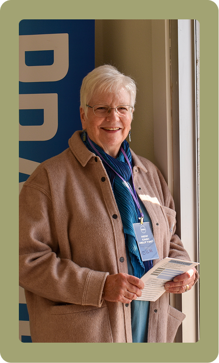 Smiling person in a tan coat standing by a doorway, holding papers and wearing a blue lanyard.