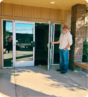 Man standing at an open glass door entrance to a building, wearing a hat and jeans.