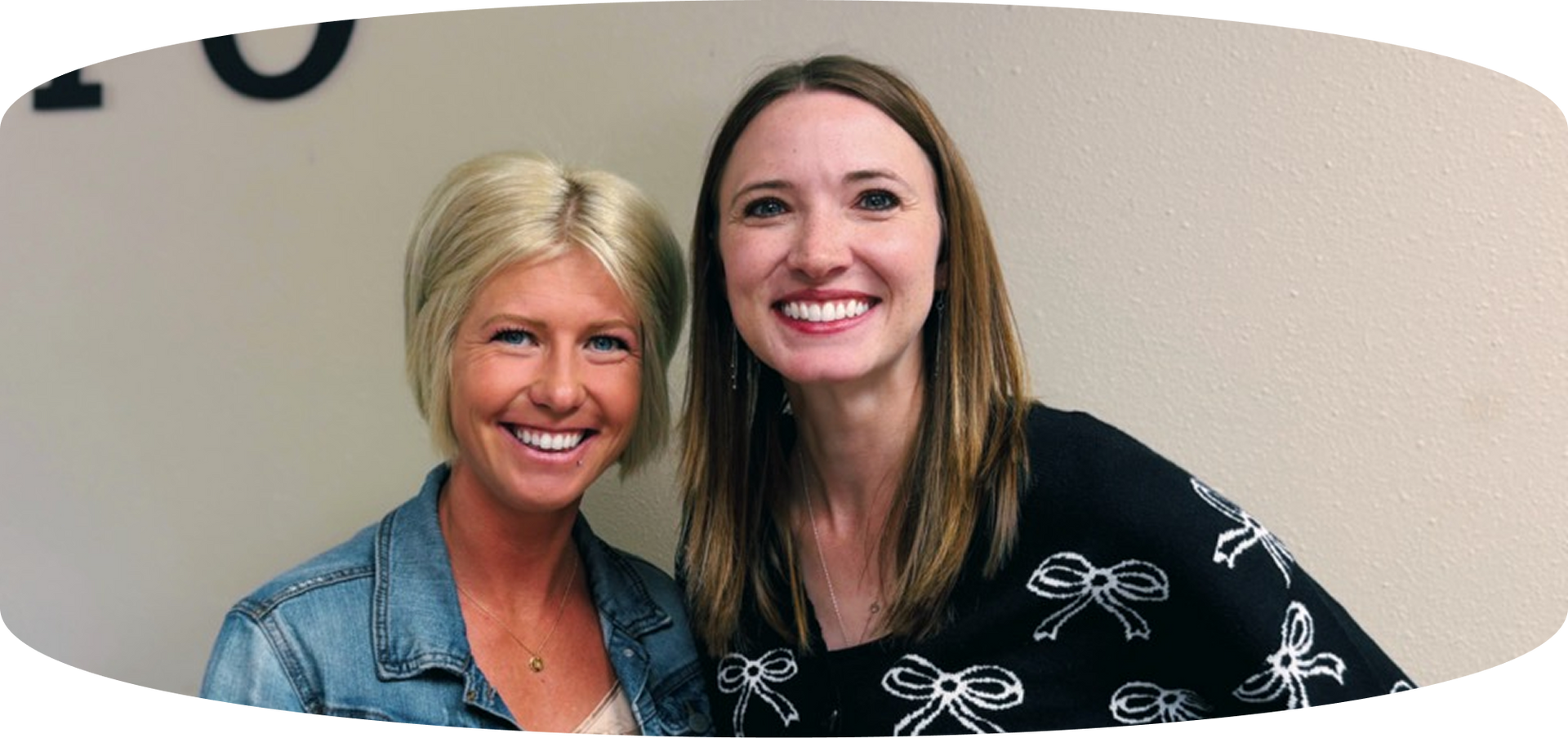 Two women smiling and posing together indoors against a beige wall.