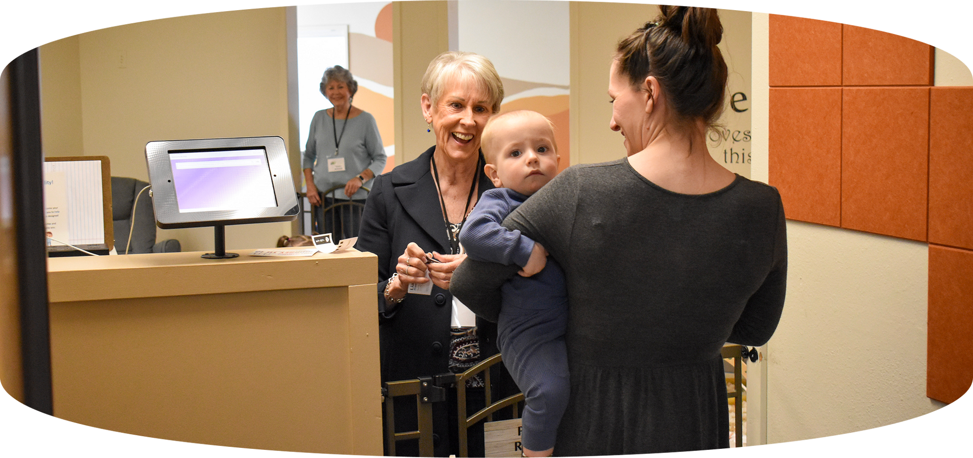 Woman with a baby at a reception desk, speaking to a smiling clerk in a clinic or office.