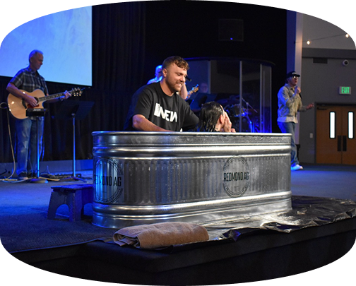 Man standing in a large metal tub on a church stage with musicians and blue lighting in the background