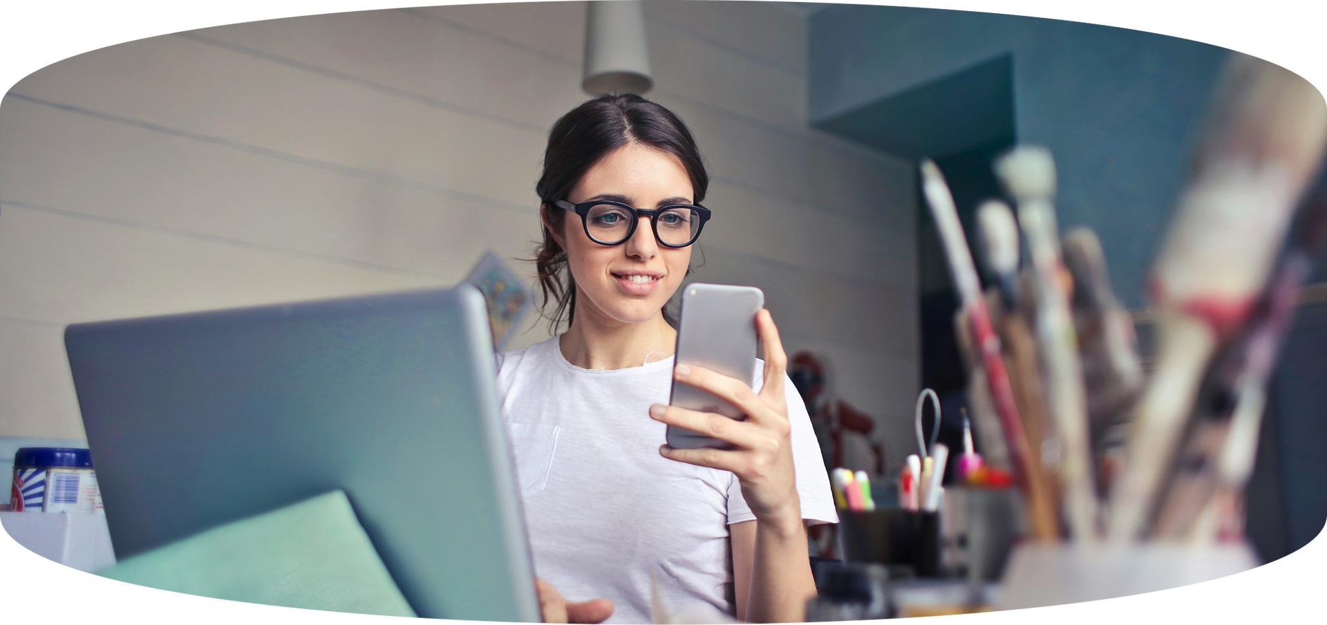 Woman with glasses smiling at a laptop while using a smartphone in a bright office setting