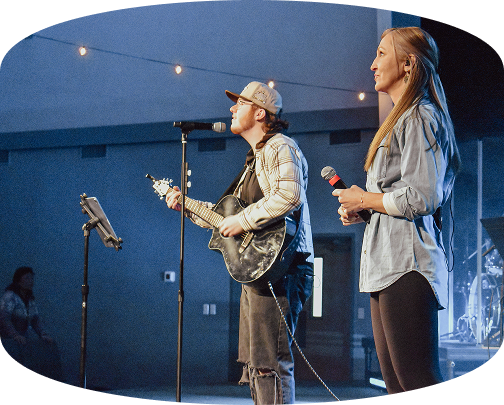 Two musicians singing on stage, one playing guitar and one holding a microphone, under blue lights