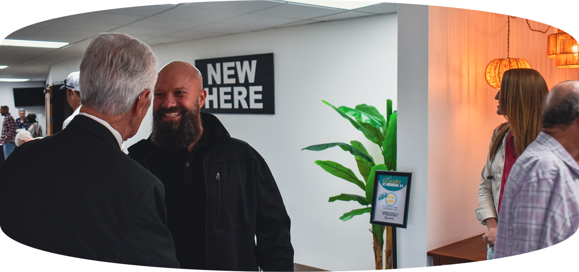 Two men talking in a lobby beside a “NEW HERE” sign and potted plant