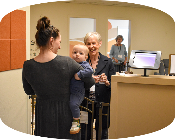 Woman holding baby at a hospital reception desk, with staff and a computer monitor nearby.