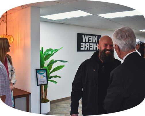 Two men chatting in a bright office lobby near a potted plant and a sign reading “NEW HERE”