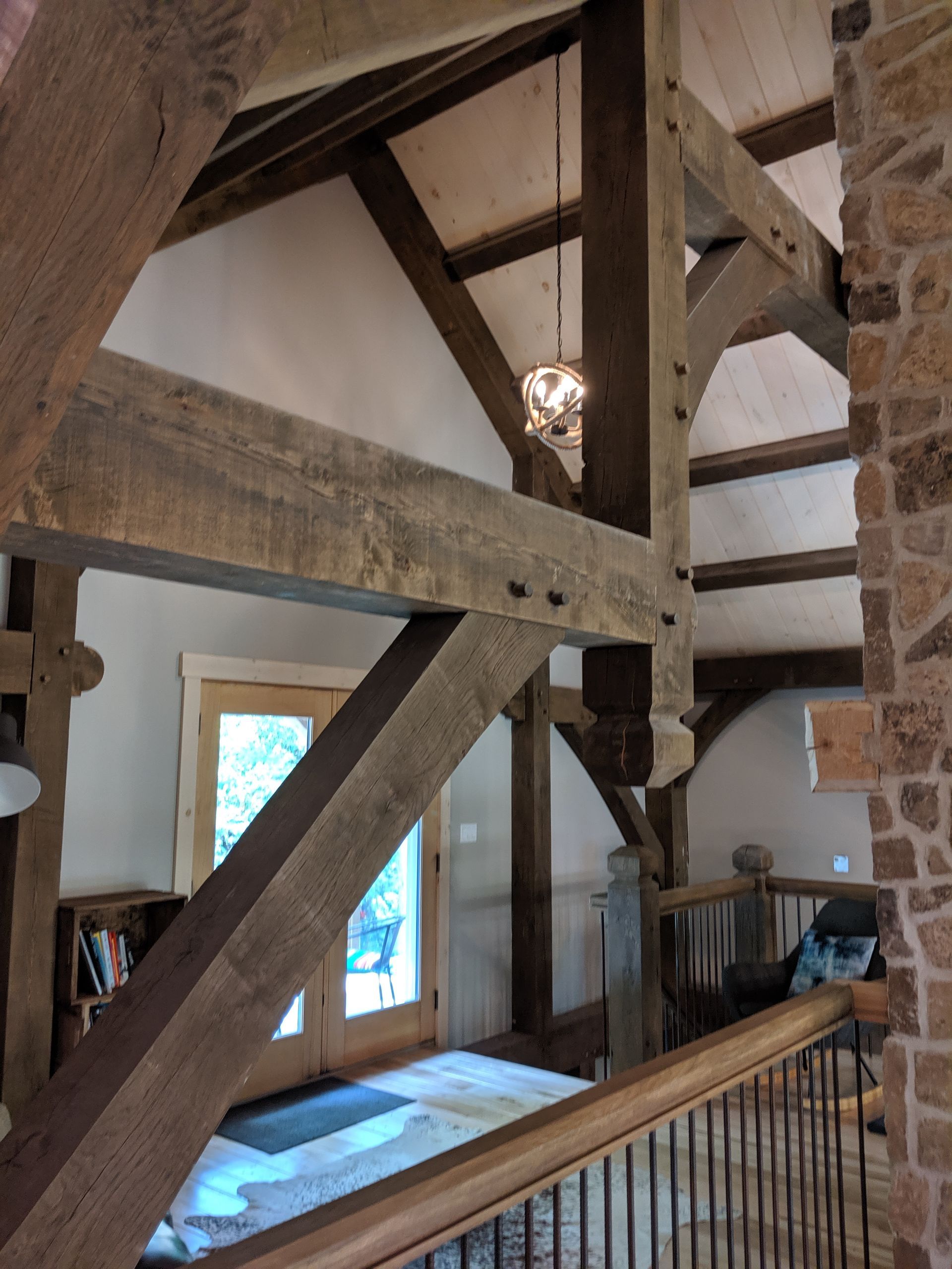 Wooden beams frame a home's interior, visible from a balcony. French doors, stone wall, and chandelier are also seen.