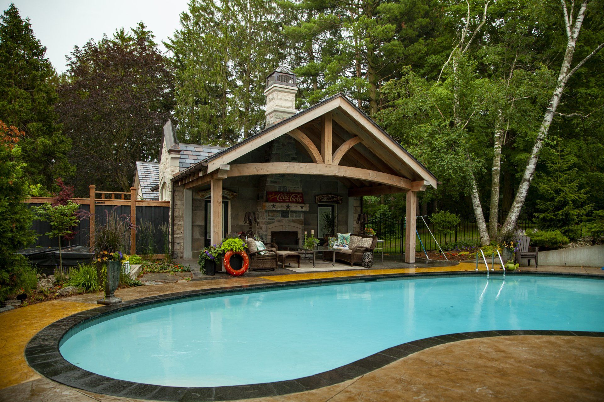 A large swimming pool is surrounded by trees and a timber frame pool house.