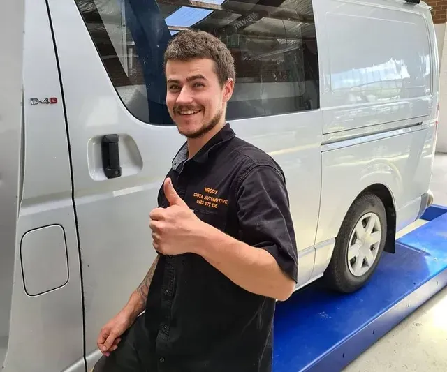 Mechanic giving thumbs up next to a silver van in a garage.