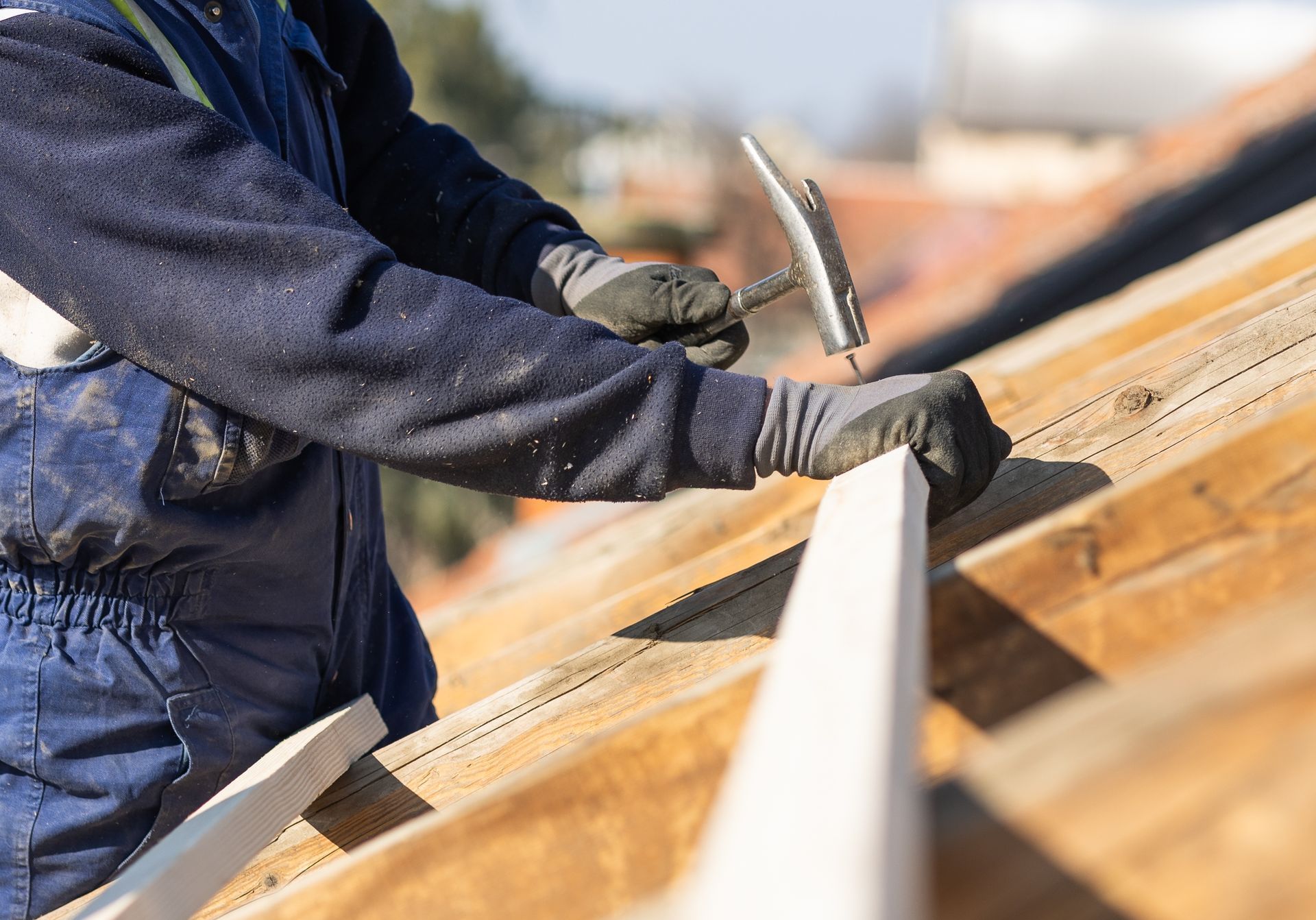 A person in blue workwear uses a hammer to attach a wooden beam to a roof structure.