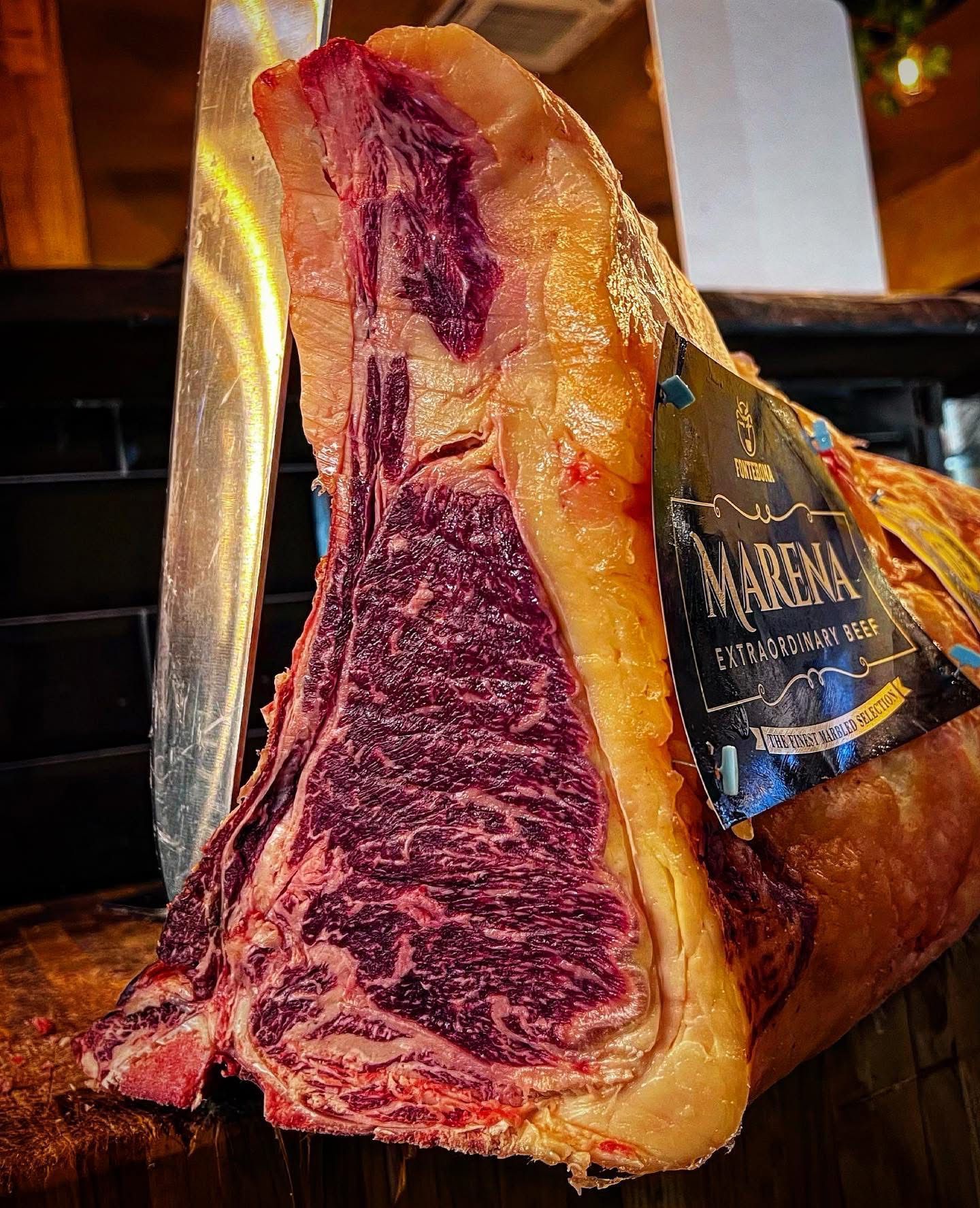 Una grande bistecca di ribeye cruda, ricca di marezzature e con osso. Accanto ad essa è posizionato un coltello.