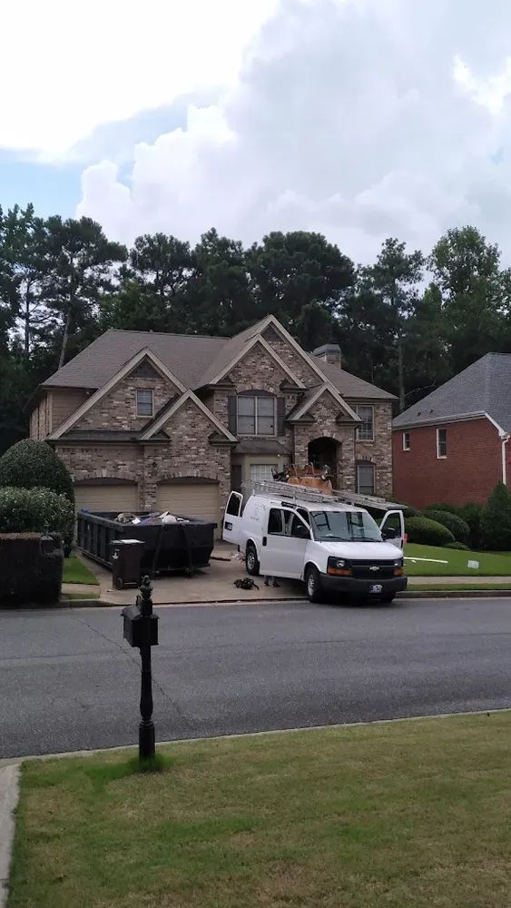 A white work van parked in front of a brick house with a dumpster in the driveway. Trees and cloudy sky in the background.