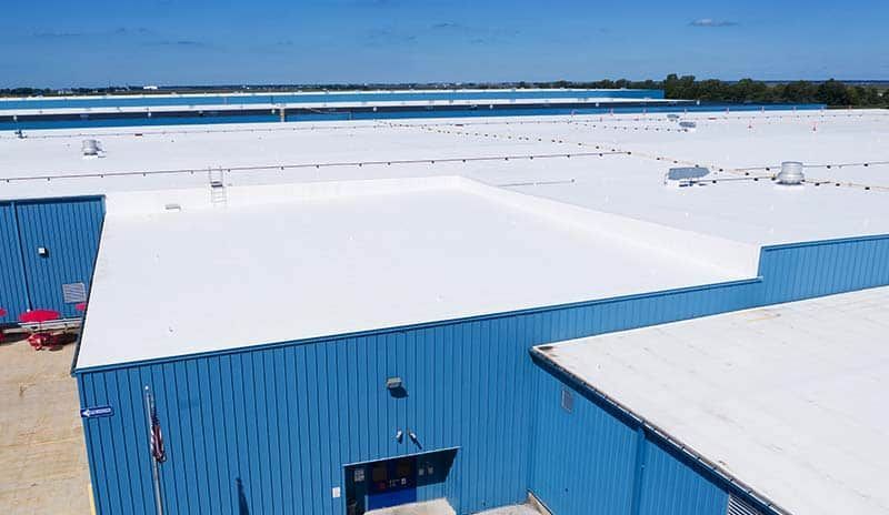Blue industrial building with white flat roof under a blue sky, near water and a bridge.