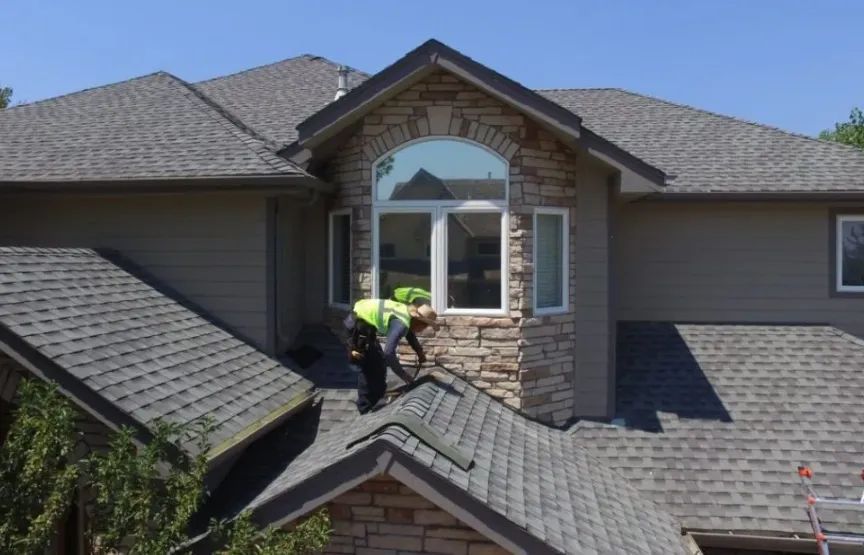 Roofer in safety gear works on gray shingled roof of a house with stone facade, under a clear blue sky.
