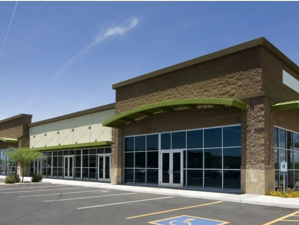 Exterior of a strip mall with large windows, tan brick facade, green awnings, and a parking lot.