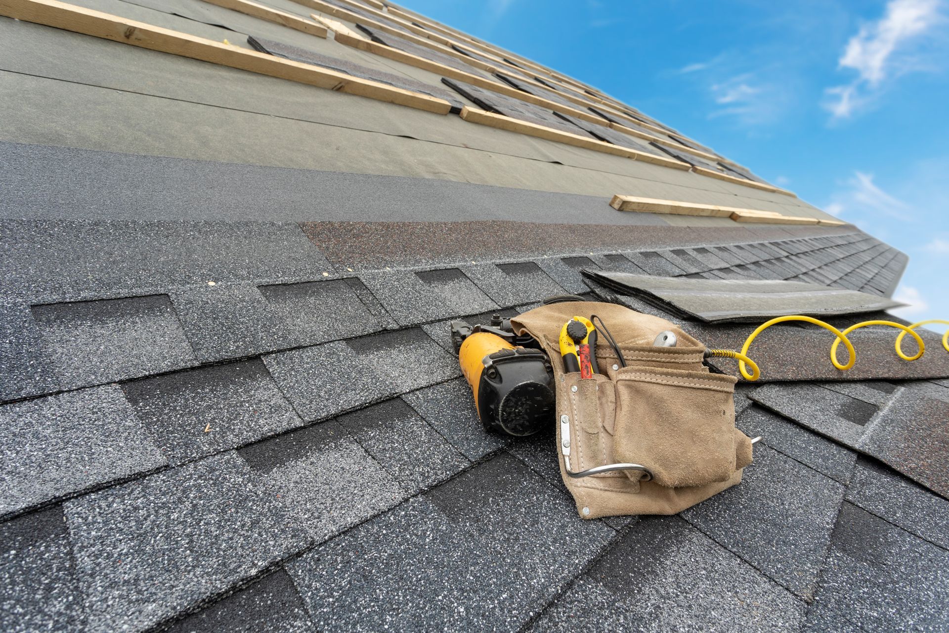 Roof being shingled; close-up shows shingles, pneumatic nailer, and tool belt on the roof against a blue sky.