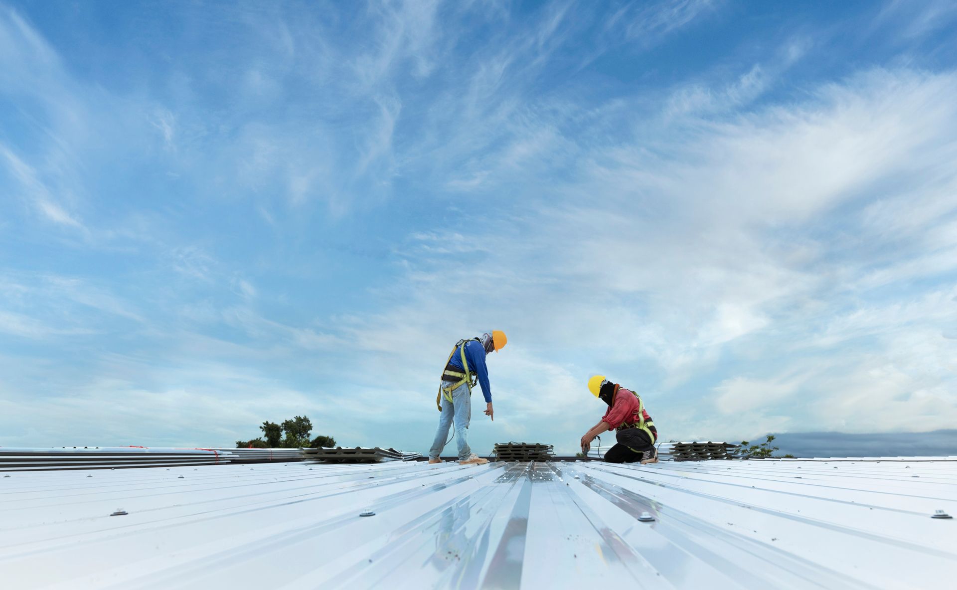 Two workers in safety harnesses on a reflective metal roof under a blue sky, installing or repairing.